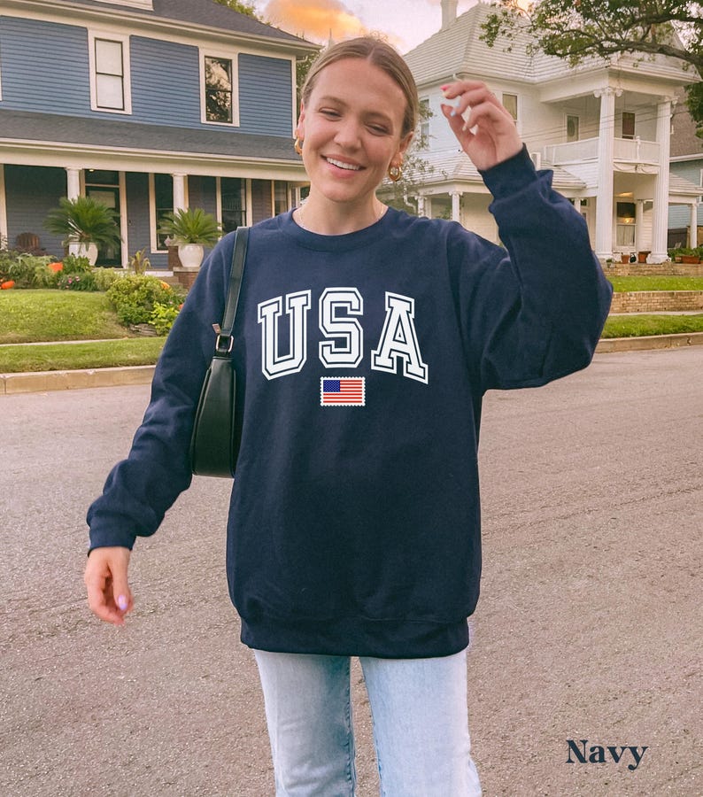 Pu&ograve; includere: Felpa blu navy con una grafica bianca "USA" e una piccola bandiera americana.