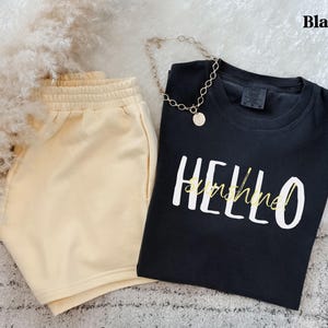 May include: A black t-shirt with the words "HELLO sunshine!" in white and yellow script, alongside beige shorts and a gold necklace. The shorts have an elasticated waistband. The word "Black" is in the top right corner.