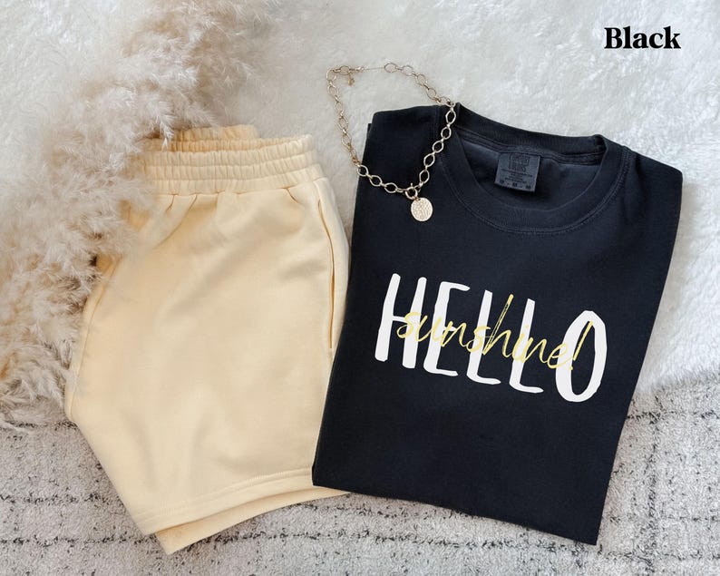 May include: A black t-shirt with the words "HELLO sunshine!" in white and yellow script, alongside beige shorts and a gold necklace. The shorts have an elasticated waistband. The word "Black" is in the top right corner.