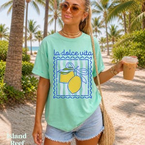 Pu&ograve; includere: T-shirt verde chiaro con la scritta "la dolce vita" e una grafica di due limoni gialli legati con un nastro blu. La maglietta ha una vestibilit&agrave; comoda.