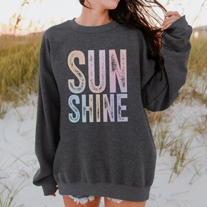 May include: Dark heather gray sweatshirt with the word "SUNSHINE" in large, pastel-colored letters. The sweatshirt is a crew neck style and is made of a soft material. The text is in a gradient of colors, including pink, blue, and yellow.