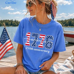 May include: A blue t-shirt with the text "1776 America 250th anniversary" in red, white, and blue. The shirt is worn with denim shorts and sunglasses. An American flag is in the background.