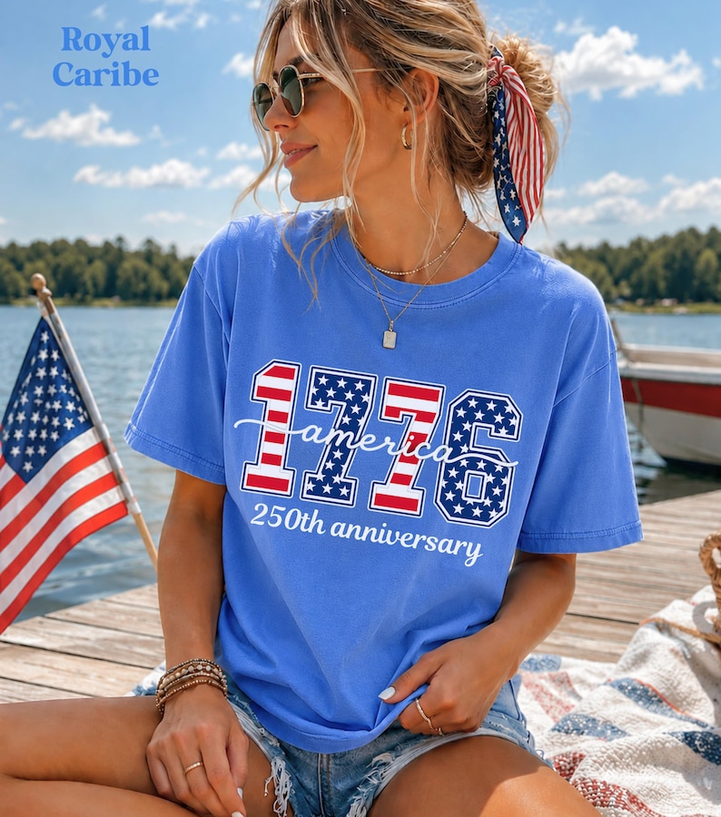 May include: A blue t-shirt with the text "1776 America 250th anniversary" in red, white, and blue. The shirt is worn with denim shorts and sunglasses. An American flag is in the background.