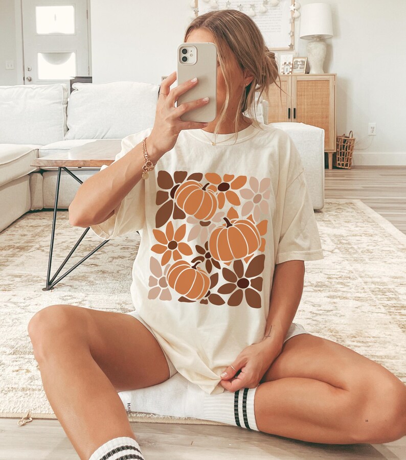 May include: Cream-colored t-shirt featuring a fall-themed design. The graphic includes three orange pumpkins and various brown and tan flowers. The shirt is a relaxed fit, suitable for casual wear.
