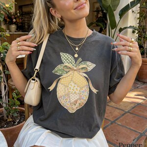 May include: Dark grey t-shirt with a patchwork lemon design, bow, and leaves. The lemon is made of floral and gingham fabrics. The word "Pepper" is visible in the bottom right corner of the image.