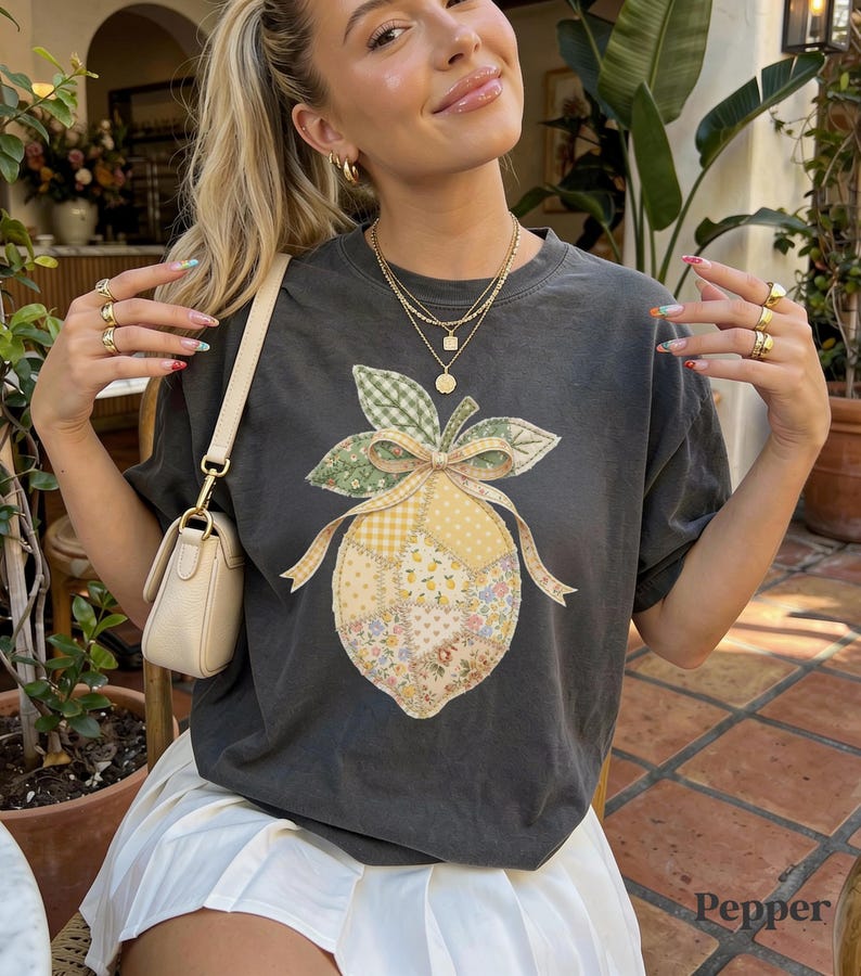 May include: Dark grey t-shirt with a patchwork lemon design, bow, and leaves. The lemon is made of floral and gingham fabrics. The word "Pepper" is visible in the bottom right corner of the image.