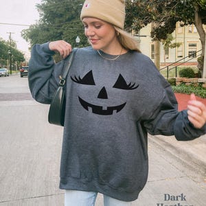 May include: A dark heather grey crewneck sweatshirt featuring a black graphic of a jack-o'-lantern face with long eyelashes. The sweatshirt is paired with a tan beanie and a black shoulder bag, showcasing a casual long-sleeve design.