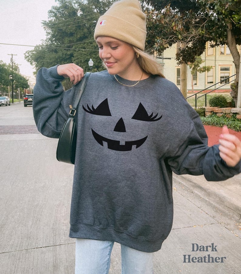 May include: A dark heather grey crewneck sweatshirt featuring a black graphic of a jack-o'-lantern face with long eyelashes. The sweatshirt is paired with a tan beanie and a black shoulder bag, showcasing a casual long-sleeve design.