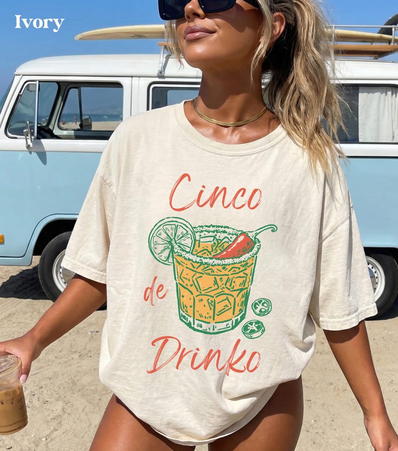 May include: Ivory-colored t-shirt with a Cinco de Drinko graphic. The design features a cocktail with a lime, chili pepper, and ice cubes. The text "Cinco de Drinko" is in red and green. The shirt is worn on a beach.