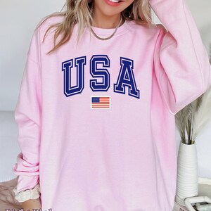 Pu&ograve; includere: Felpa rosa chiaro con la scritta "USA" in lettere blu scuro con contorno bianco e una piccola bandiera americana sotto.