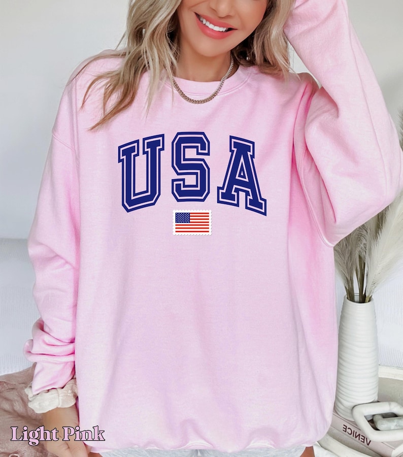 Pu&ograve; includere: Felpa rosa chiaro con la scritta "USA" in lettere blu scuro con contorno bianco e una piccola bandiera americana sotto.