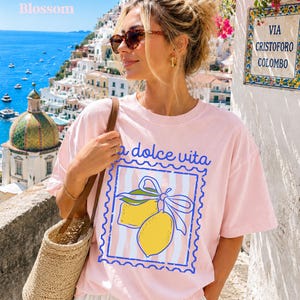 Pu&ograve; includere: T-shirt rosa chiaro con la scritta "la dolce vita" e un disegno di limone in un riquadro blu. La maglietta ha uno sfondo a righe. La parola "Blossom" &egrave; nell'angolo in alto a sinistra.