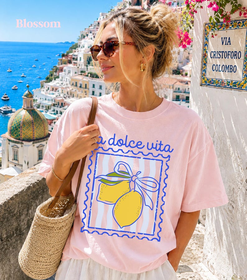 Pu&ograve; includere: T-shirt rosa chiaro con la scritta "la dolce vita" e un disegno di limone in un riquadro blu. La maglietta ha uno sfondo a righe. La parola "Blossom" &egrave; nell'angolo in alto a sinistra.