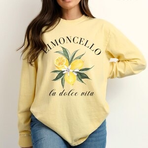 May include: Butter-colored long-sleeve shirt with a "Limoncello" design. The design features lemons, leaves, and flowers, with the phrase "la dolce vita" below. The shirt is made of a soft material and is suitable for casual wear.