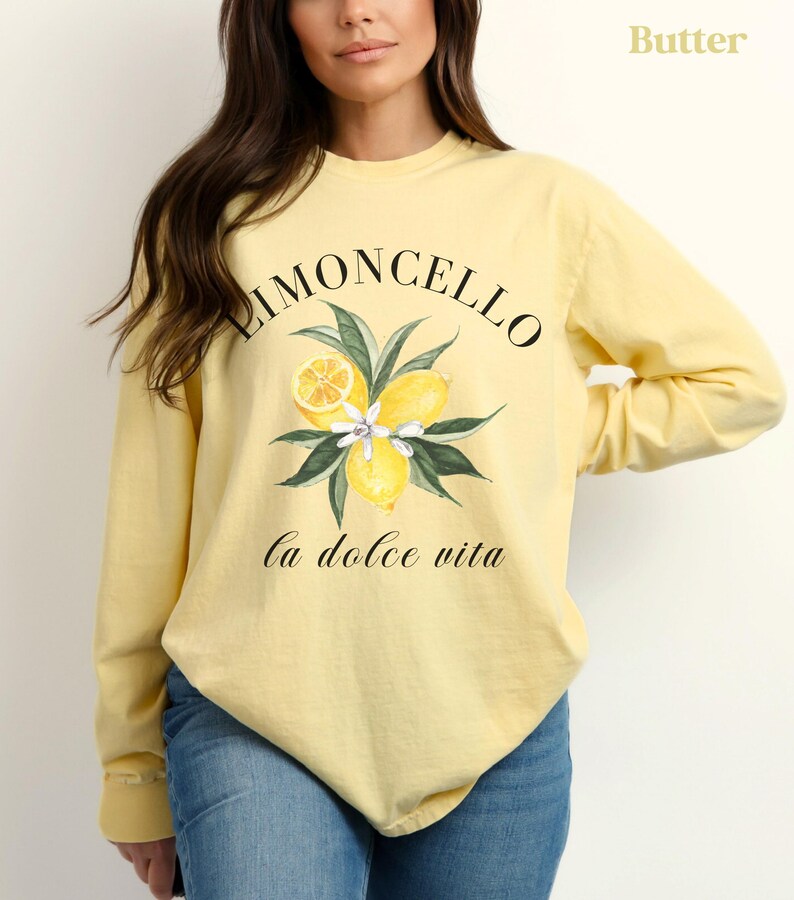 May include: Butter-colored long-sleeve shirt with a "Limoncello" design. The design features lemons, leaves, and flowers, with the phrase "la dolce vita" below. The shirt is made of a soft material and is suitable for casual wear.
