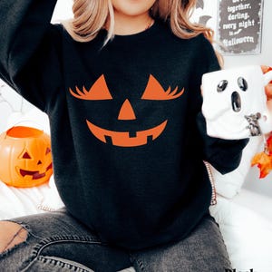 May include: A black long-sleeve sweatshirt featuring a large orange graphic of a jack-o'-lantern face with long eyelashes. The design includes triangular eyes with lashes, a small triangular nose, and a wide, jagged, smiling mouth.