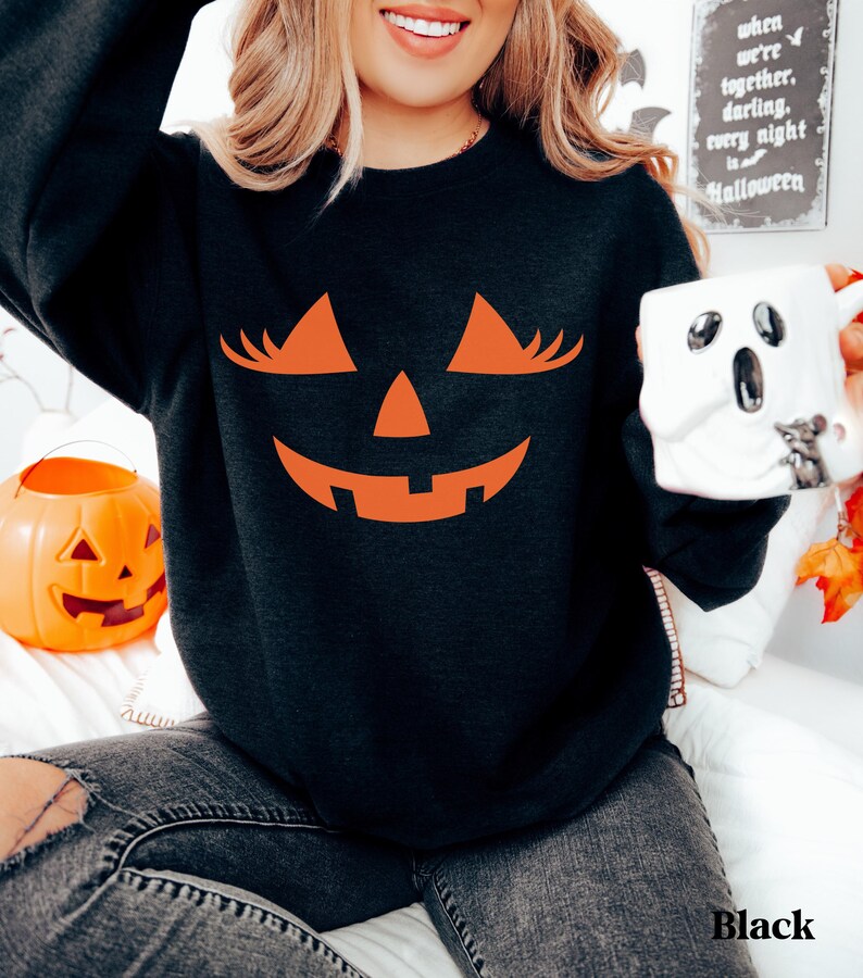 May include: A black long-sleeve sweatshirt featuring a large orange graphic of a jack-o'-lantern face with long eyelashes. The design includes triangular eyes with lashes, a small triangular nose, and a wide, jagged, smiling mouth.