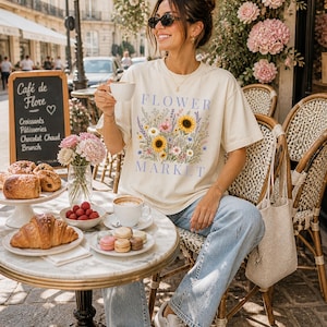 May include: A cream-colored short-sleeve t-shirt featuring a graphic print of a colorful wildflower bouquet with sunflowers and the text "FLOWER MARKET" in blue lettering. The shirt has a crew neckline and a relaxed fit, made of cotton fabric.
