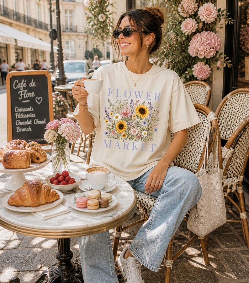 May include: A cream-colored short-sleeve t-shirt featuring a graphic print of a colorful wildflower bouquet with sunflowers and the text "FLOWER MARKET" in blue lettering. The shirt has a crew neckline and a relaxed fit, made of cotton fabric.