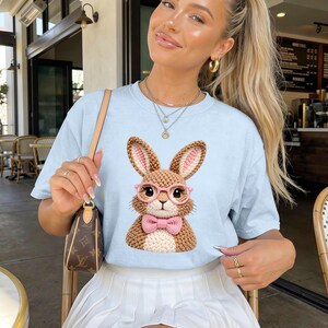 May include: Light blue t-shirt with a graphic of a brown bunny wearing pink glasses and a pink bow tie. The word "Chambray" is printed at the top left. The shirt is worn with a white pleated skirt.
