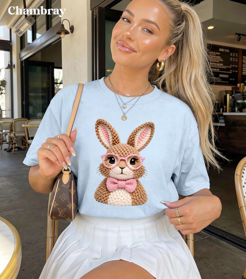May include: Light blue t-shirt with a graphic of a brown bunny wearing pink glasses and a pink bow tie. The word "Chambray" is printed at the top left. The shirt is worn with a white pleated skirt.