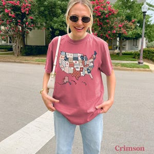 May include: A crimson red t-shirt with a patchwork design of the United States of America. The design includes the words "Brave", "Loved", "Free", and "United".