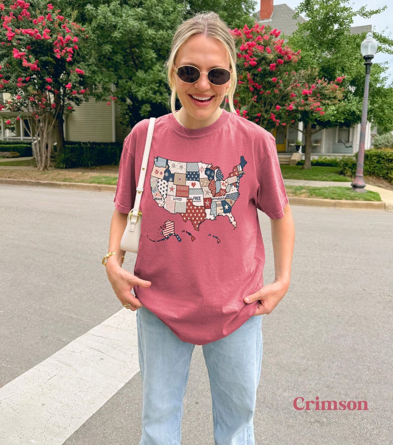 May include: A crimson red t-shirt with a patchwork design of the United States of America. The design includes the words "Brave", "Loved", "Free", and "United".