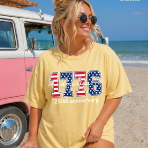 May include: A yellow t-shirt with the text "1776 America 250th anniversary" in red, white, and blue. The shirt is worn by a person with sunglasses, standing on a beach with a vintage van in the background.