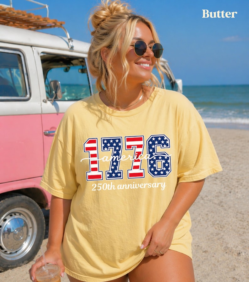 May include: A yellow t-shirt with the text "1776 America 250th anniversary" in red, white, and blue. The shirt is worn by a person with sunglasses, standing on a beach with a vintage van in the background.