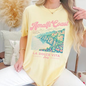 May include: A yellow t-shirt with a graphic of the Amalfi Coast, Italy. The graphic features a colorful illustration of the coastline with buildings and flowers. The text "Amalfi Coast" is in pink and the text "La Dolce Vita Italy" is in black.