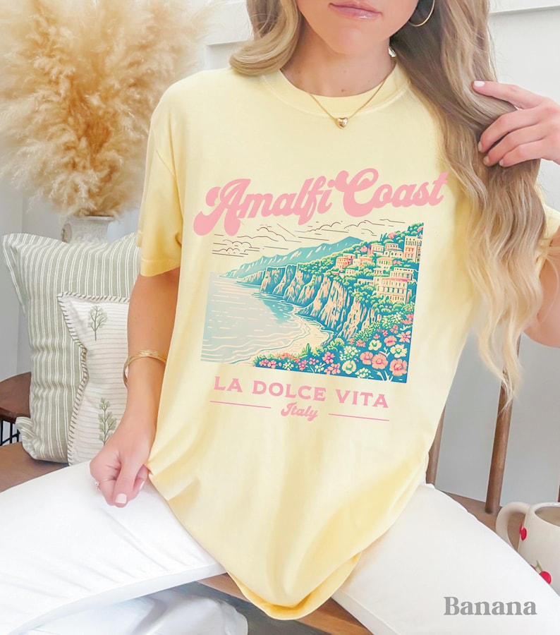 May include: A yellow t-shirt with a graphic of the Amalfi Coast, Italy. The graphic features a colorful illustration of the coastline with buildings and flowers. The text "Amalfi Coast" is in pink and the text "La Dolce Vita Italy" is in black.