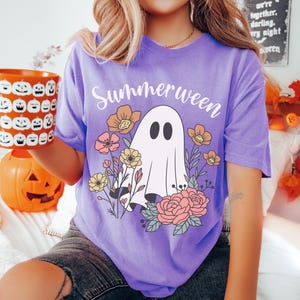 May include: A violet t-shirt with the word "Summerween" above a ghost graphic surrounded by flowers. The ghost has two black oval eyes. The shirt is worn by a person holding a pumpkin mug.