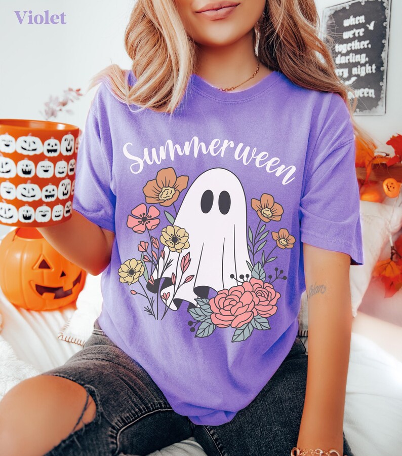 May include: A violet t-shirt with the word "Summerween" above a ghost graphic surrounded by flowers. The ghost has two black oval eyes. The shirt is worn by a person holding a pumpkin mug.