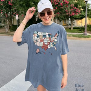 May include: A blue jean t-shirt with a patchwork design of the United States map. The map is made up of different colored squares with words like "brave", "loved", "free", and "victorious" written on them. The shirt is oversized and has a relaxed fit.