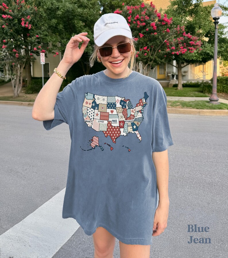 May include: A blue jean t-shirt with a patchwork design of the United States map. The map is made up of different colored squares with words like "brave", "loved", "free", and "victorious" written on them. The shirt is oversized and has a relaxed fit.