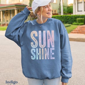 May include: Indigo blue sweatshirt with the word "SUNSHINE" in a gradient of pastel colors. The sweatshirt is a crew neck style. The text "Indigo Blue" is at the bottom left of the image. The person is wearing a white baseball cap.