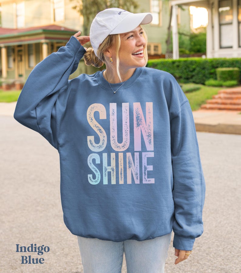 May include: Indigo blue sweatshirt with the word "SUNSHINE" in a gradient of pastel colors. The sweatshirt is a crew neck style. The text "Indigo Blue" is at the bottom left of the image. The person is wearing a white baseball cap.