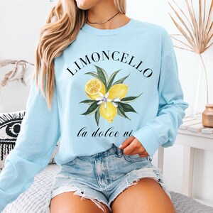 May include: Light blue long-sleeve shirt with a watercolor illustration of lemons, leaves, and flowers. The word "LIMONCELLO" is arched above the illustration, with "la dolce vi" below. The shirt is a casual style.