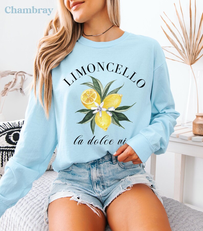 May include: Light blue long-sleeve shirt with a watercolor illustration of lemons, leaves, and flowers. The word "LIMONCELLO" is arched above the illustration, with "la dolce vi" below. The shirt is a casual style.
