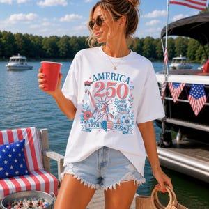 K&ouml;nnte beinhalten: Wei&szlig;es T-Shirt mit einer Grafik zum 250. Jahrestag Amerikas, mit der Freiheitsstatue, floralen Akzenten und den Jahren 1776 und 2026. Die Person tr&auml;gt Jeansshorts und eine Sonnenbrille und h&auml;lt einen roten Becher und eine Korbtasche.