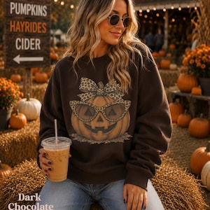 Op de afbeelding: Een donker chocoladebruin sweatshirt met lange mouwen en een centrale afbeelding van een pompoen met een luipaardprint bril en een bijpassende strik, gecombineerd met een lichtblauwe spijkerbroek en een doorzichtige plastic beker.