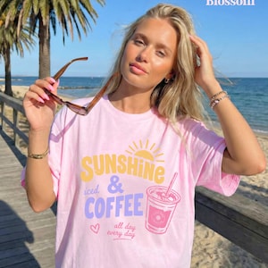 Puede incluir: Camiseta rosa claro con el texto "Sunshine & Iced Coffee all day every day" en amarillo, morado y rosa. La camiseta presenta un gr&aacute;fico de un sol y un caf&eacute; helado. La persona lleva gafas de sol.
