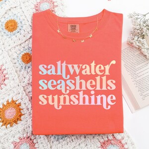 sunshine beach vacation shirt that says saltwater seashells sunshine in a fun summer print on a Comfort Colors shirt in the color bright salmon orange
