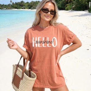 May include: A terracotta t-shirt with the text "HELLO sunshine!" in white. The image shows a person wearing sunglasses and holding a straw tote bag with a brown leather detail. The background is a beach with turquoise water and palm trees.