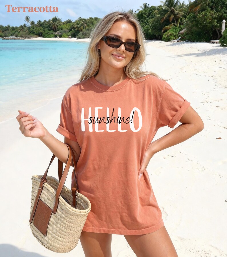 May include: A terracotta t-shirt with the text "HELLO sunshine!" in white. The image shows a person wearing sunglasses and holding a straw tote bag with a brown leather detail. The background is a beach with turquoise water and palm trees.