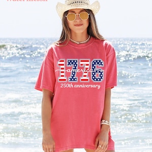 May include: A coral-coloured t-shirt featuring the text "1776 America 250th anniversary" in red, white, and blue. The shirt is worn with a straw hat and sunglasses. The background is a beach with ocean waves.
