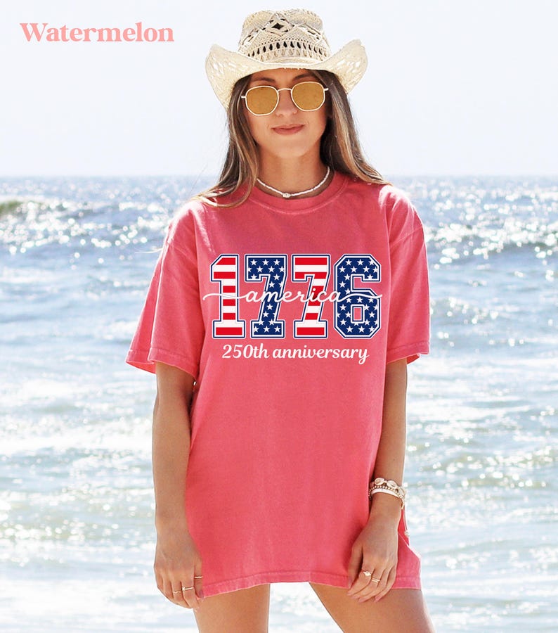 May include: A coral-coloured t-shirt featuring the text "1776 America 250th anniversary" in red, white, and blue. The shirt is worn with a straw hat and sunglasses. The background is a beach with ocean waves.