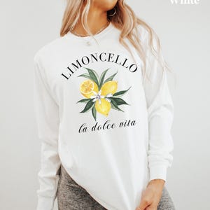 May include: White long-sleeve shirt with a watercolor design featuring lemons, leaves, and the text "Limoncello" and "la dolce vita". The shirt is a casual style, perfect for everyday wear.