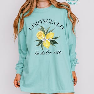 May include: A long-sleeve, chalky mint green shirt featuring the word "Limoncello" above a watercolor illustration of lemons and leaves. Below the illustration, the phrase "la dolce vita" is written in cursive. The shirt is a comfortable, casual style.