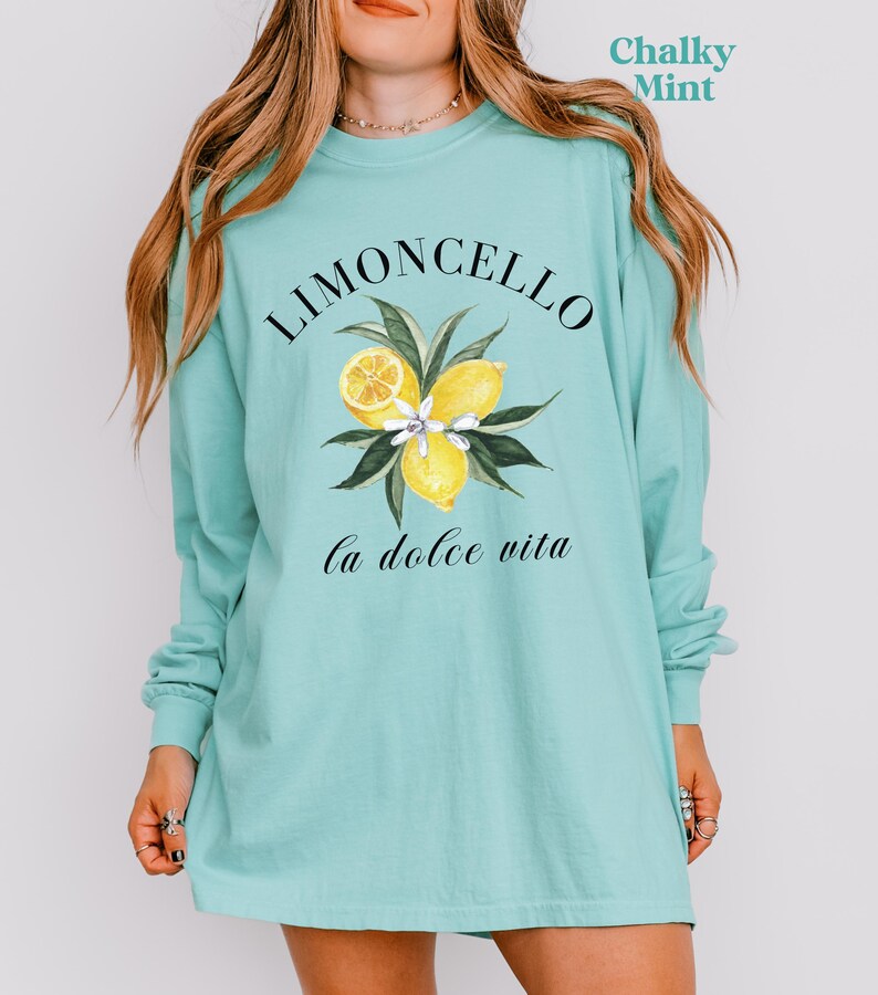 May include: A long-sleeve, chalky mint green shirt featuring the word "Limoncello" above a watercolor illustration of lemons and leaves. Below the illustration, the phrase "la dolce vita" is written in cursive. The shirt is a comfortable, casual style.